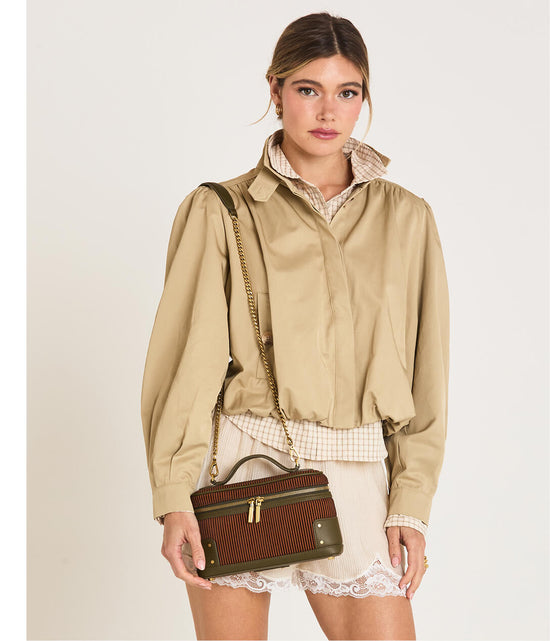 A model wearing a beige cropped utility jacket over a checkered shirt and white lace-trimmed shorts, carrying a brown and olive green striped cosmetic bag with a gold chain strap.