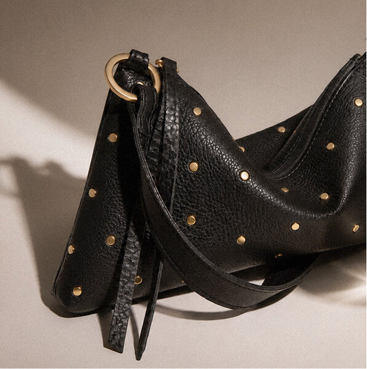 Black pebbled leather shoulder bag with gold-tone studs, photographed on a warm beige studio background.