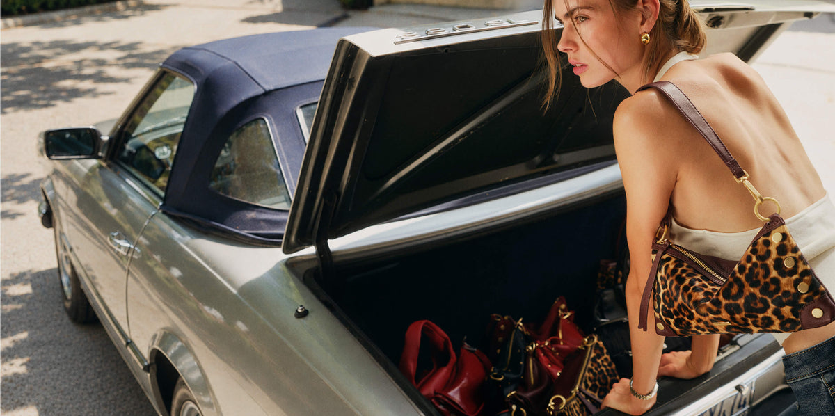 A woman with her back turned leans into the open trunk of a vintage silver convertible car, placing a leopard-print shoulder bag next to other handbags. She is wearing a white tank top and jeans.