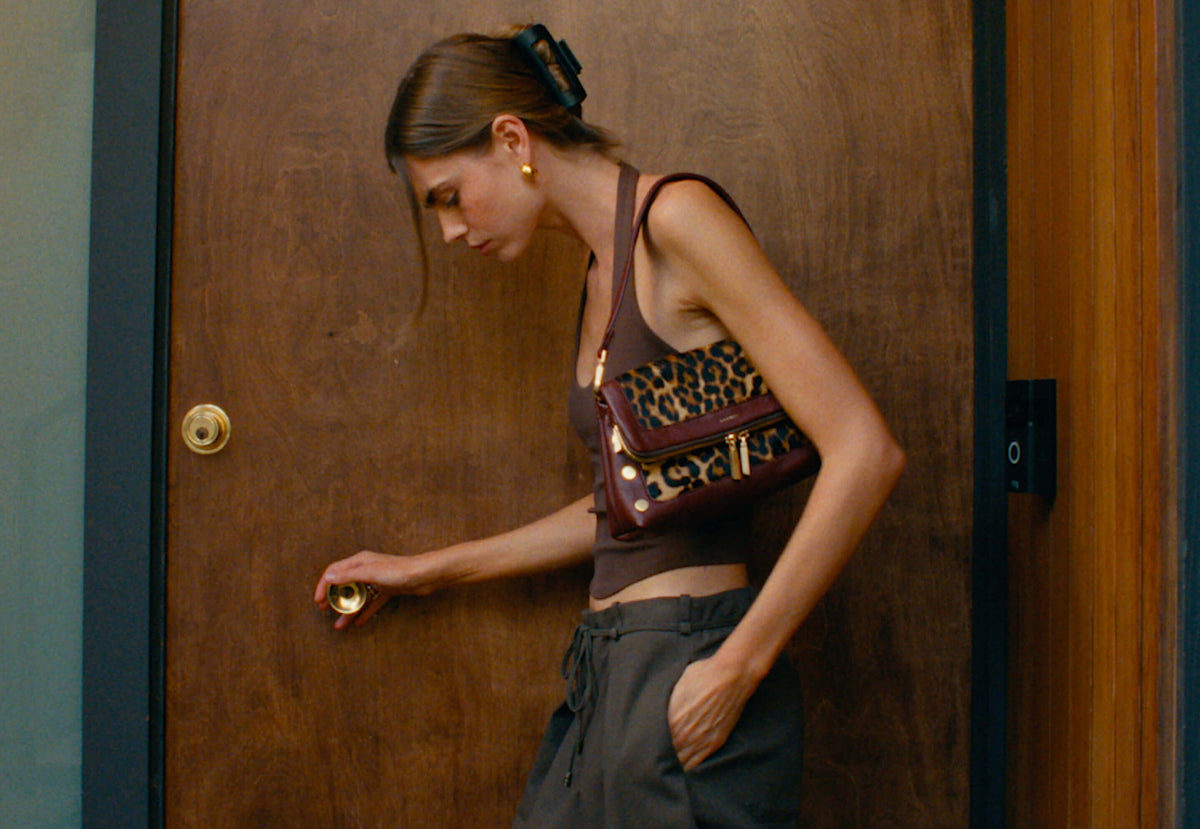 A woman in a brown tank top and dark pants stands beside a wooden door, holding a leopard-print shoulder bag with a burgundy base and gold hardware.