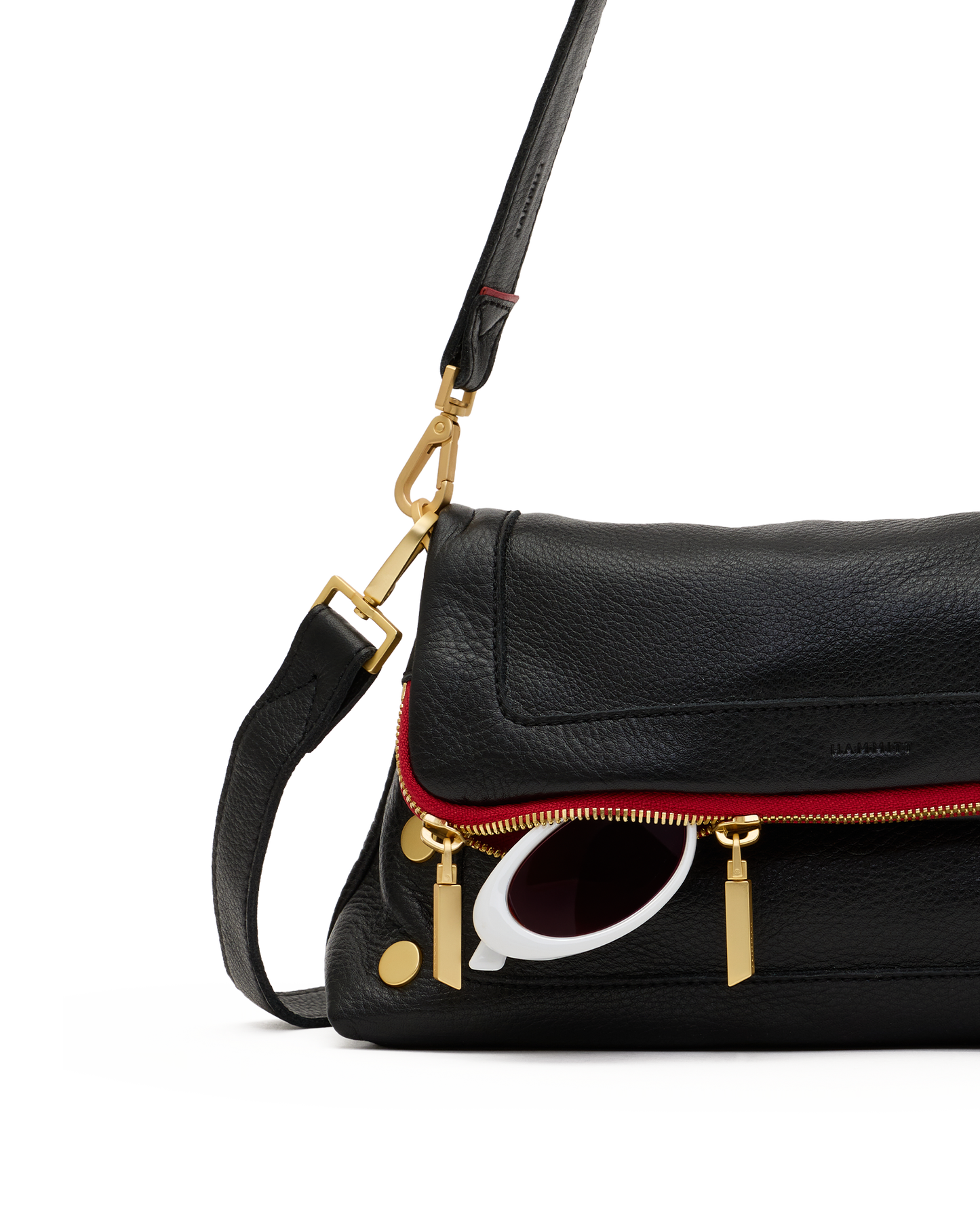 A close-up of a black pebbled leather shoulder bag with a red piping accent along the front zipper, gold-tone hardware, and a white-framed sunglasses charm, on a white background.