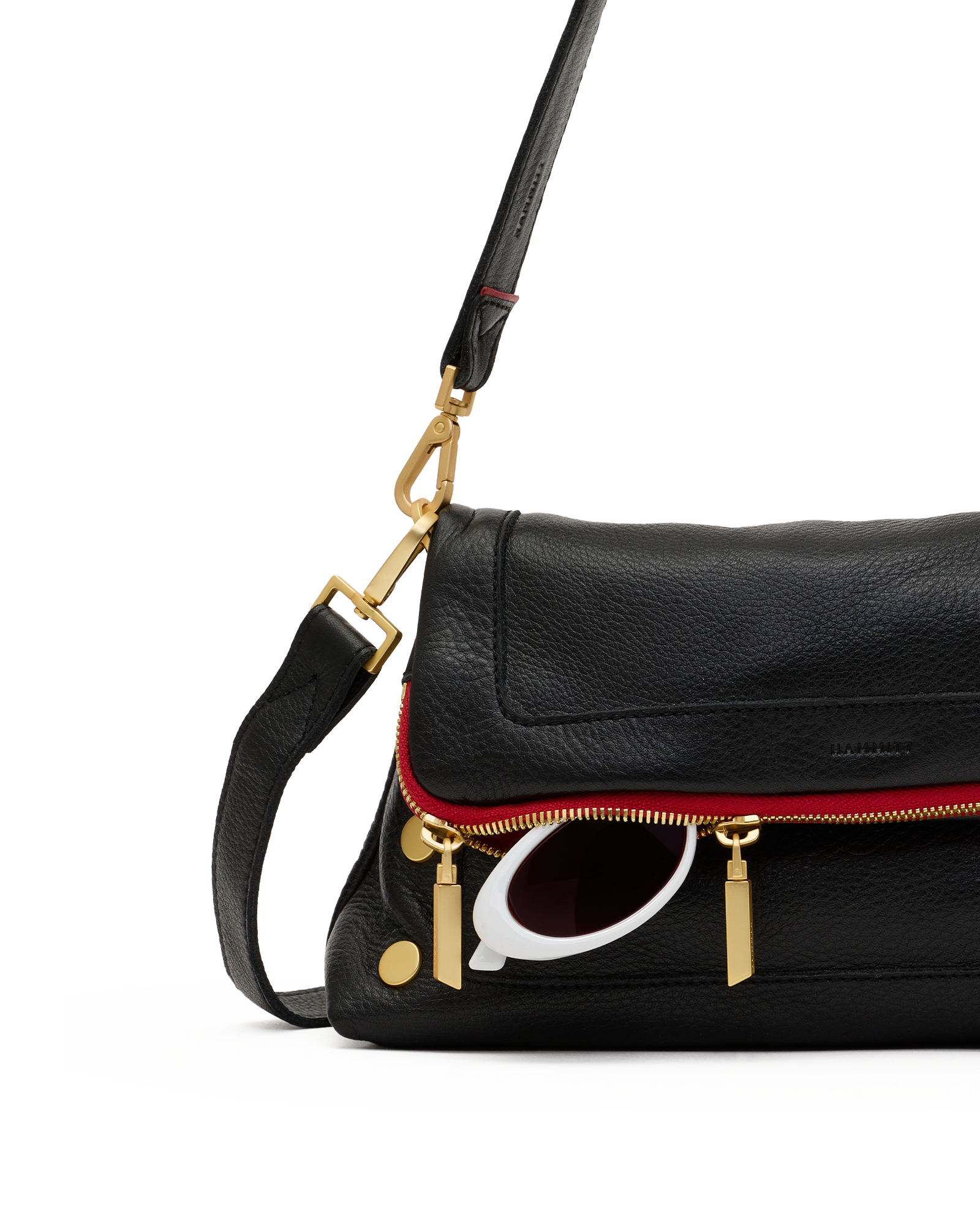 A close-up of a black pebbled leather shoulder bag with a red piping accent along the front zipper, gold-tone hardware, and a white-framed sunglasses charm, on a white background.