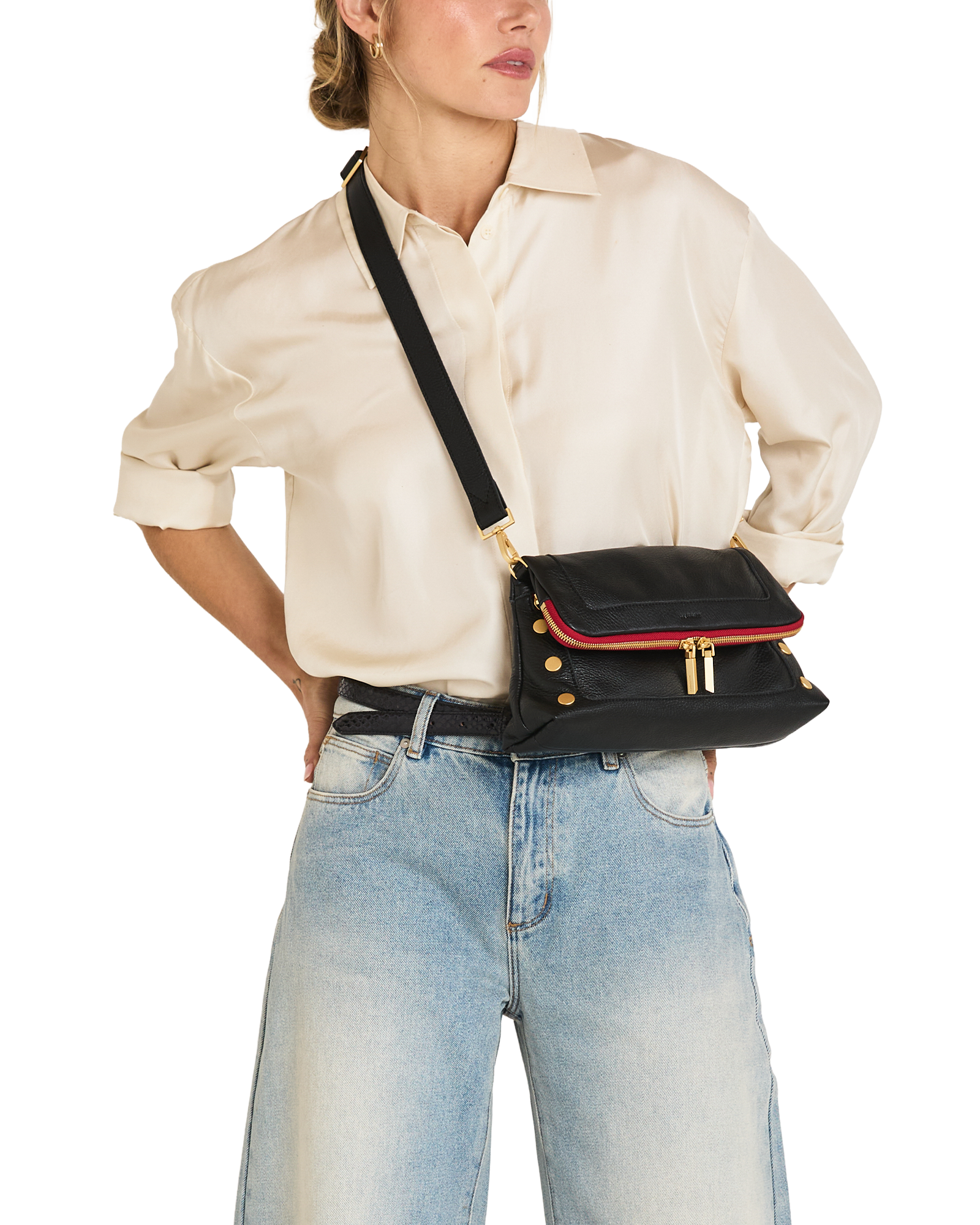 A model wearing a cream satin shirt and light blue jeans carries a black leather crossbody bag with a red leather trim and gold-tone hardware.