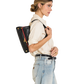A woman in a cream shirt and light blue jeans shown from the side, wearing a black leather shoulder bag with gold hardware and a red accent stripe against a plain white background.