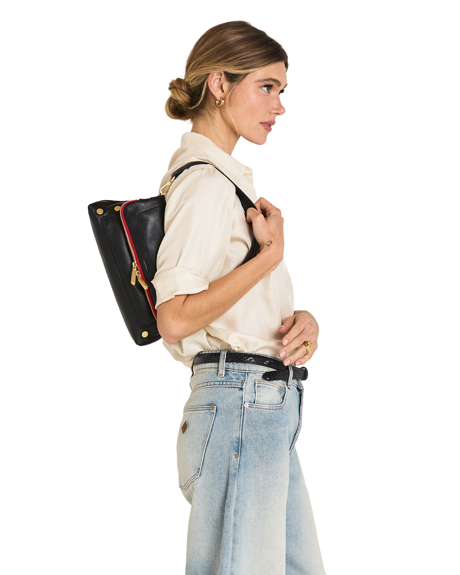 A woman in a cream shirt and light blue jeans shown from the side, wearing a black leather shoulder bag with gold hardware and a red accent stripe against a plain white background.