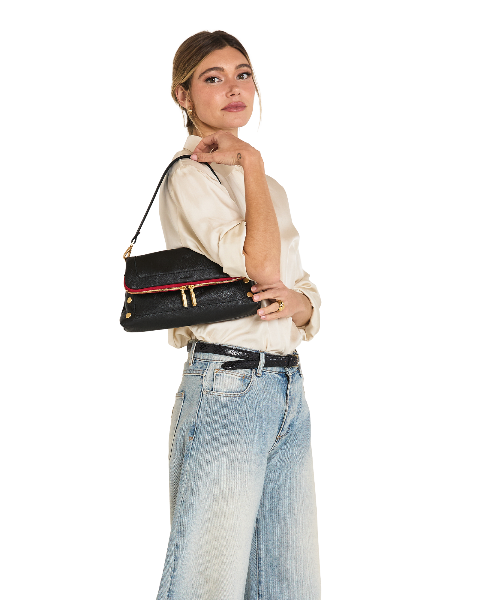 A woman wearing a cream blouse and light blue jeans carries a black shoulder bag with a red trim and gold hardware against a plain white background.