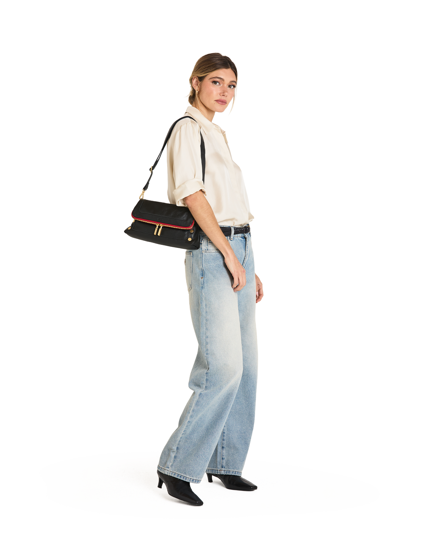A woman modeling a black shoulder bag with red and gold trim, styled with a cream short-sleeve blouse, light-wash wide-leg jeans, and black pointed-toe heels against a plain white background.