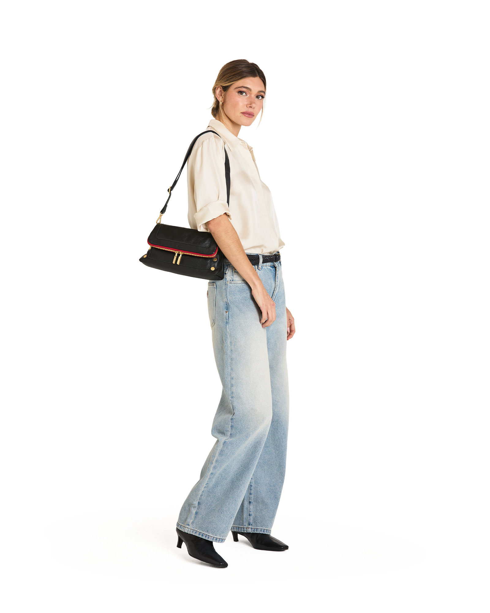 A woman modeling a black shoulder bag with red and gold trim, styled with a cream short-sleeve blouse, light-wash wide-leg jeans, and black pointed-toe heels against a plain white background.