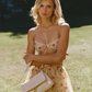 A woman standing outdoors on a grassy lawn wearing a floral bralette and matching skirt, holding a light beige crossbody bag.