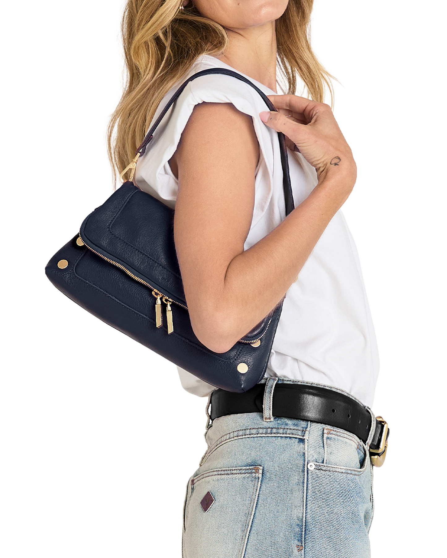 A woman wearing a white sleeveless top and light blue jeans carries a navy blue leather shoulder bag with gold-tone hardware against a plain white background.