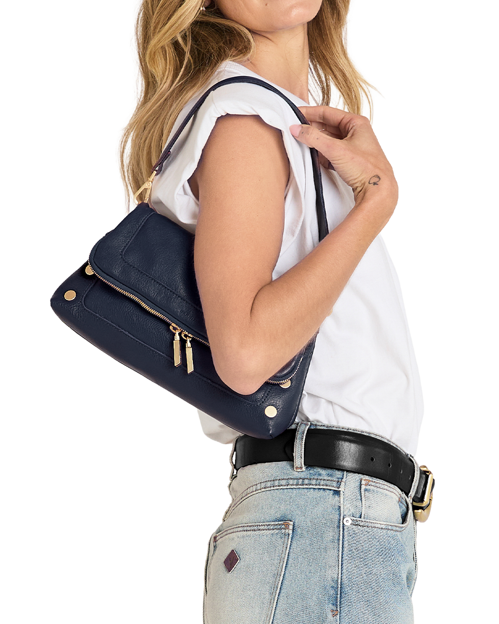 A woman wearing a white sleeveless top and light blue jeans carries a navy blue leather shoulder bag with gold-tone hardware against a plain white background.