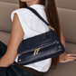 A woman wearing a white sleeveless top and dark jeans carries a navy blue leather shoulder bag with gold-tone hardware and a matching shoulder strap.
