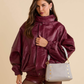 A woman models an oversized, shiny burgundy faux-leather jacket with a high collar, paired with matching burgundy shorts and a light gray shoulder bag with gold studs, against a solid beige background.