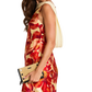 A woman wearing a sleeveless, knee-length dress with a bold red, orange, and yellow floral print, holding a beige clutch bag.