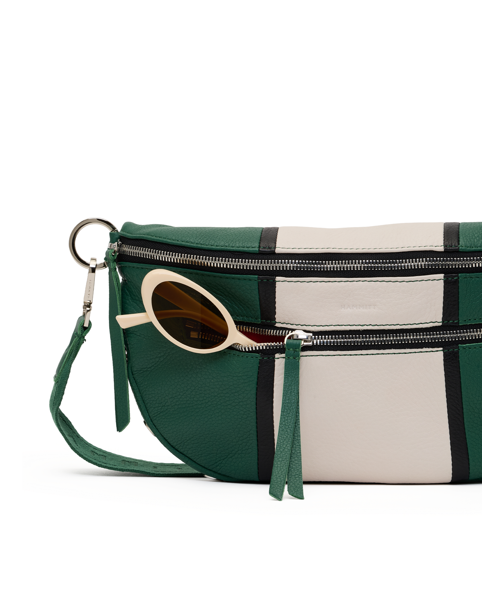 A green and cream leather crossbody bag with a geometric stripe pattern, featuring silver zippers, a green wristlet strap, and a pair of round sunglasses tucked into the front pocket.