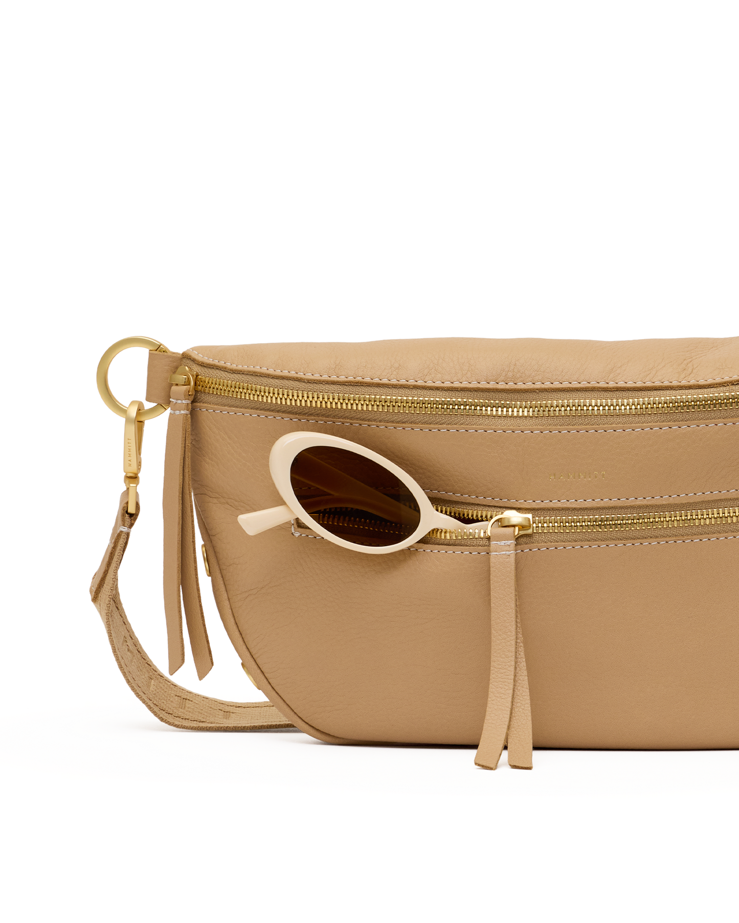 A close-up of a beige leather belt bag with gold-tone hardware, featuring a front zip pocket with a tassel zipper pull and a pair of round sunglasses resting in the pocket.