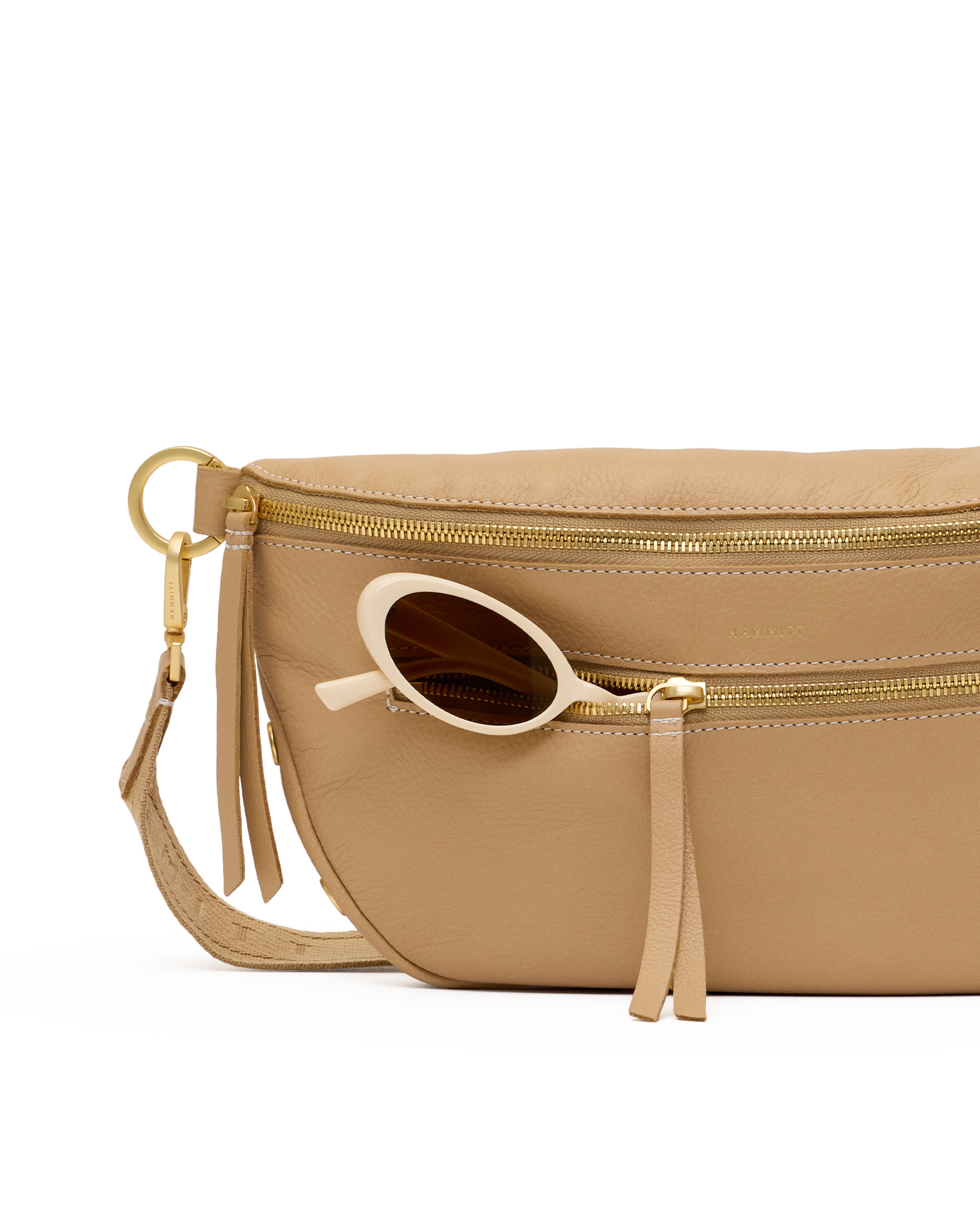 A close-up of a beige leather belt bag with gold-tone hardware, featuring a front zip pocket with a tassel zipper pull and a pair of round sunglasses resting in the pocket.