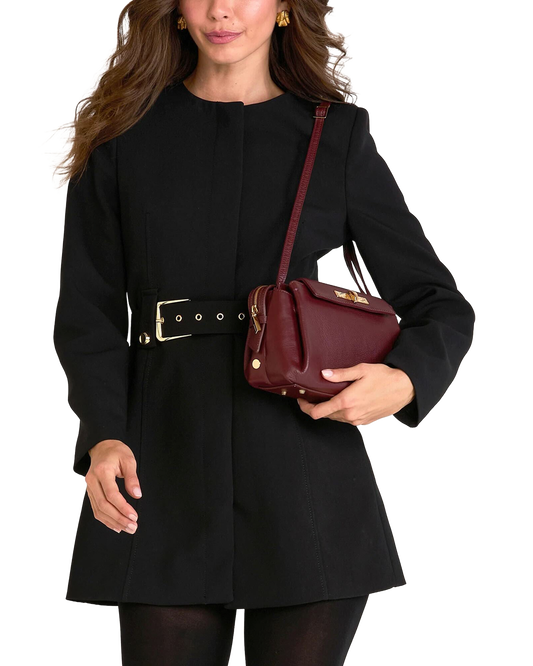 A woman wearing a black long-sleeve coat with a wide belt and holding a burgundy leather shoulder bag, shown against a plain white background.