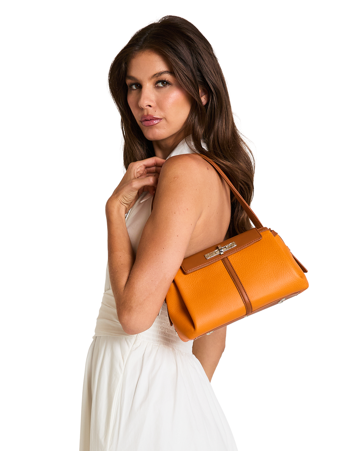 A woman in a white sleeveless dress carries a structured orange-brown leather handbag over her shoulder, photographed against a plain white background.