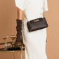 A model wearing a white dress and a long brown glove carries a dark brown textured leather crossbody bag with a silver-tone chain strap and a decorative front clasp, set against a warm beige background with a gold bar cart holding cocktails.