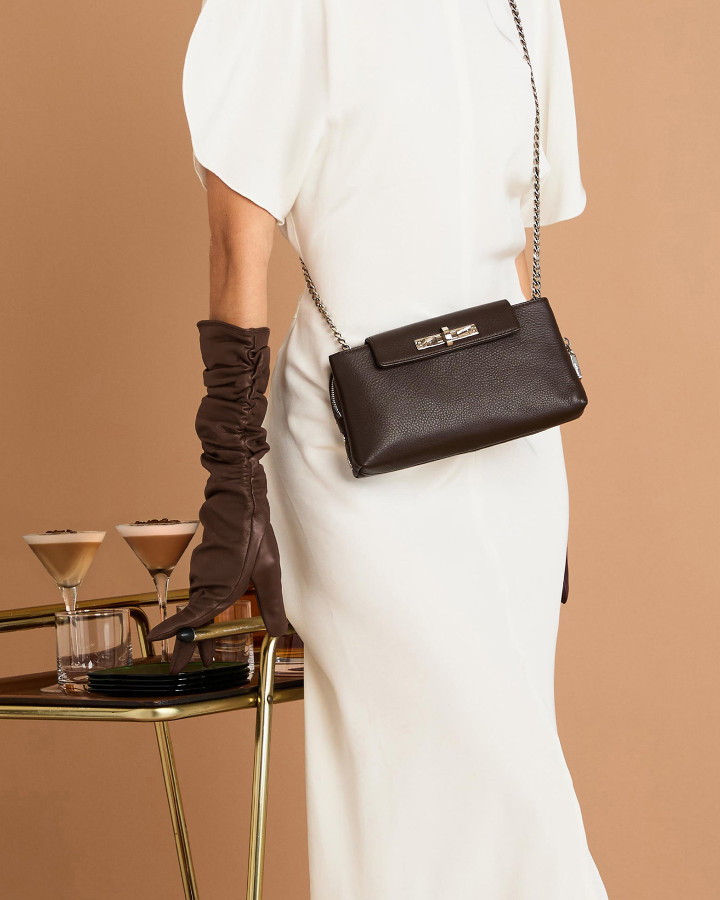 A model wearing a white dress and a long brown glove carries a dark brown textured leather crossbody bag with a silver-tone chain strap and a decorative front clasp, set against a warm beige background with a gold bar cart holding cocktails.