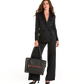 A woman modeling a black satin blazer and wide-leg trousers, holding a black leather tote bag with gold studs and a red zipper detail, against a solid white background.