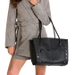 A model wearing a gray plaid belted blazer and matching shorts carries a large black leather tote bag with silver studs and a front zip pocket against a white background.