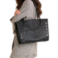 A woman wearing a gray plaid blazer and matching skirt carries a large black leather tote bag with silver-tone studs and a front zip pocket against a plain white background.
