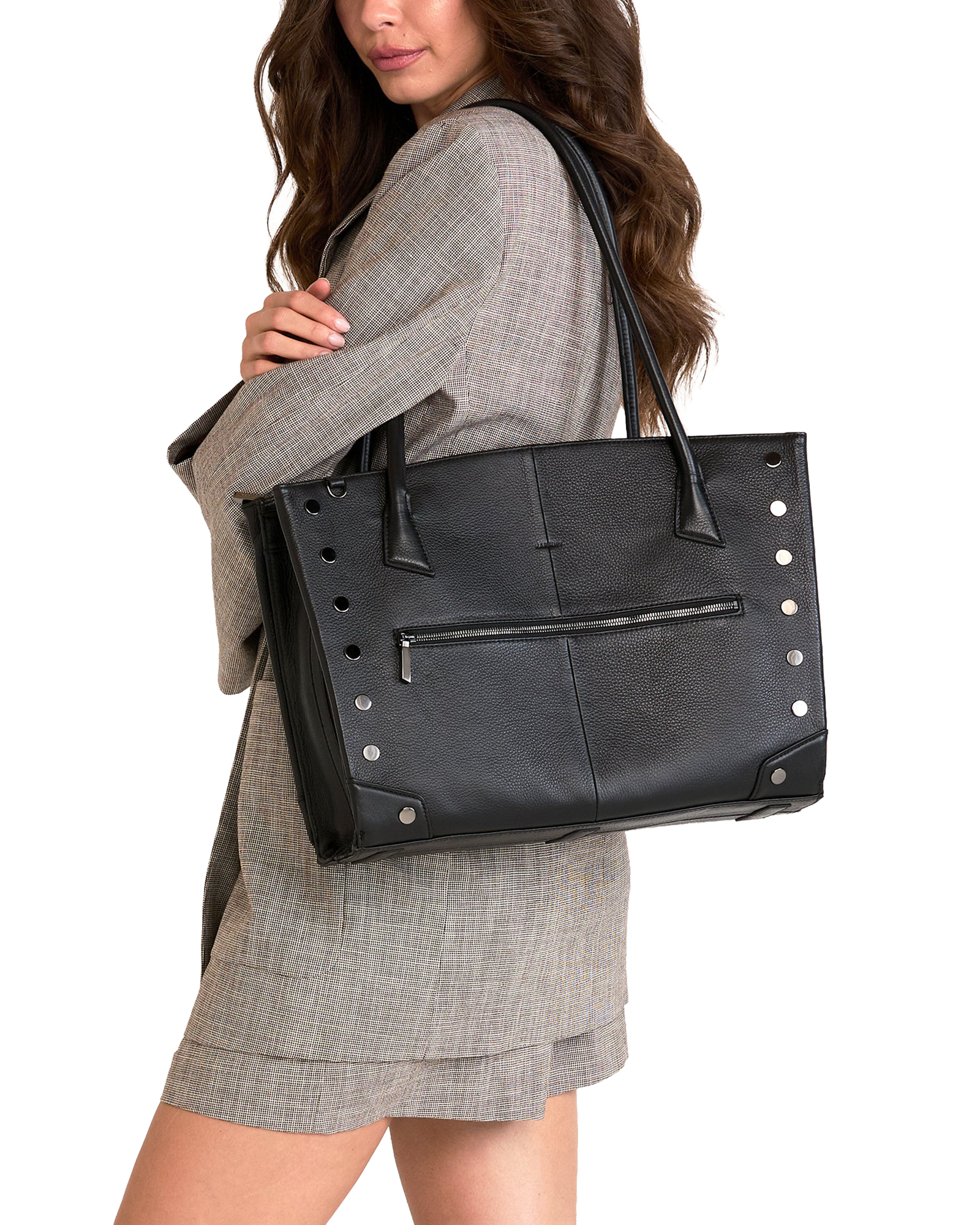 A woman wearing a gray plaid blazer and matching skirt carries a large black leather tote bag with silver-tone studs and a front zip pocket against a plain white background.