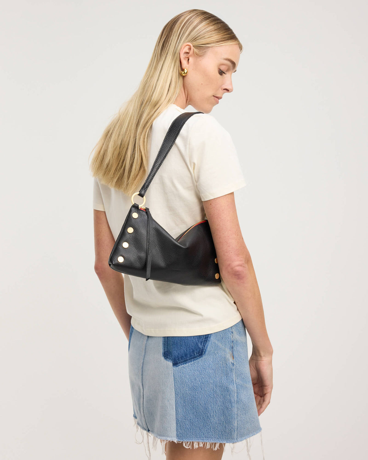 A blonde woman wearing a cream t-shirt and a denim patchwork mini skirt, shown from a side-back angle while wearing a black leather shoulder bag with gold-tone studs and a strap.