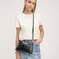 A woman modeling a black faux-leather crossbody bag with gold-tone studs and a gold buckle, styled with a cream t-shirt and denim skirt against a light gray studio background.