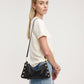 A woman in a white t-shirt and denim skirt models a black leather shoulder bag with gold studs and hardware against a plain light gray background.