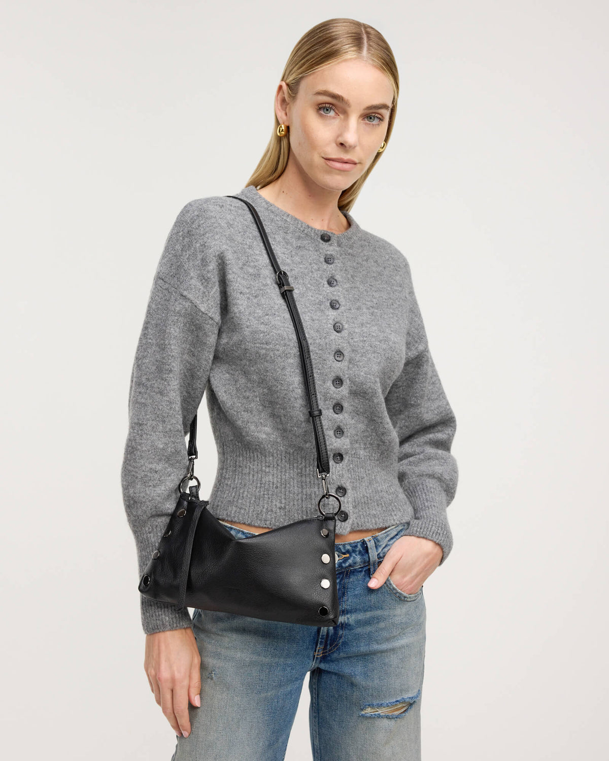 A woman wearing a gray button-front knit sweater and blue jeans, carrying a black leather shoulder bag with silver-tone hardware against a light gray studio background.