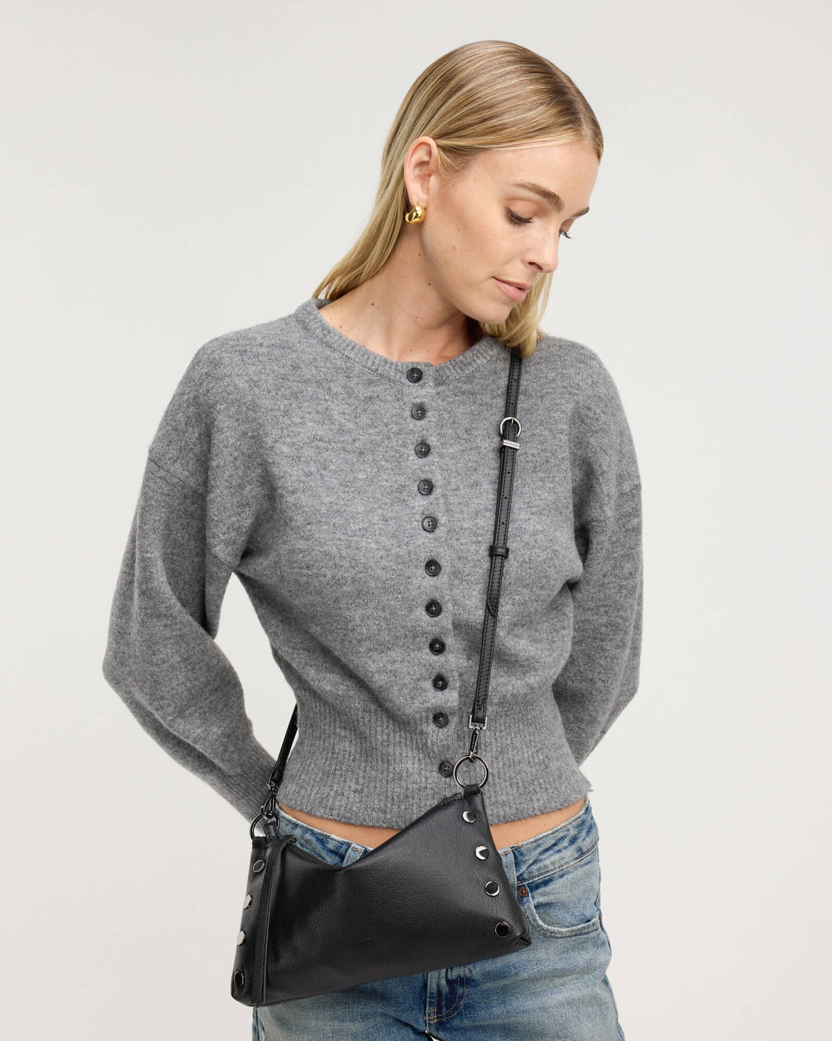 A woman wearing a gray long-sleeve button-front knit sweater and blue jeans, carrying a black crossbody bag with silver-tone hardware against a plain light gray studio background.