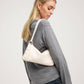 A blonde woman wearing a gray knit sweater and blue jeans, shown from a side-back angle while wearing a cream-colored slouchy shoulder bag with gold-tone hardware and decorative gold buttons.