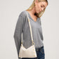 A blonde woman wearing a gray long-sleeve V-neck sweater and blue jeans, shown from the waist up. She is wearing a cream-colored shoulder bag with gold-tone hardware and a matching shoulder strap, photographed against a neutral light gray studio background.