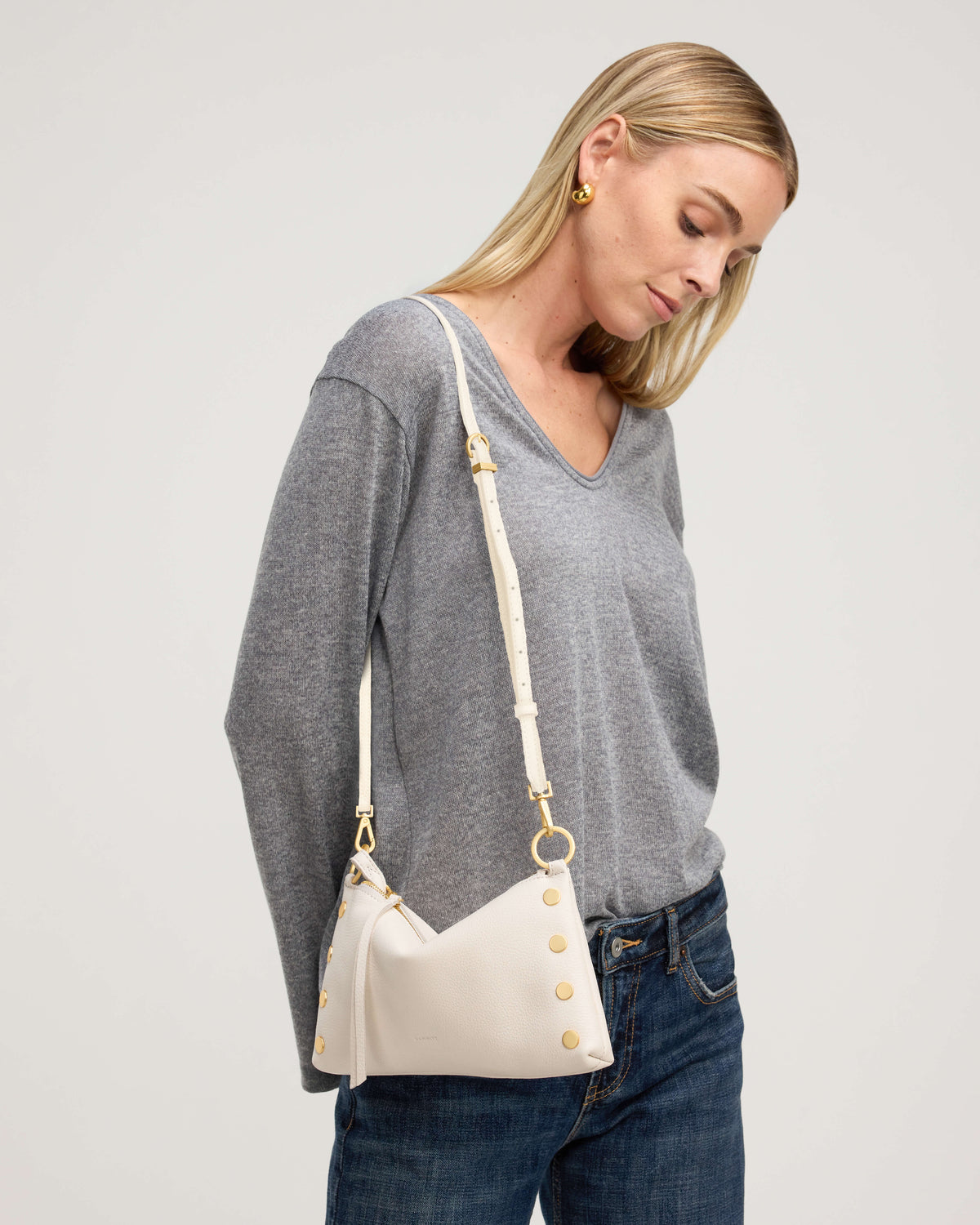 A blonde woman wearing a gray long-sleeve V-neck sweater and blue jeans, shown from the waist up. She is wearing a cream-colored shoulder bag with gold-tone hardware and a matching shoulder strap, photographed against a neutral light gray studio background.