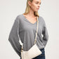 A woman wearing a gray long-sleeve V-neck sweater and dark blue jeans, carrying a cream-colored leather shoulder bag with gold hardware and a matching strap.