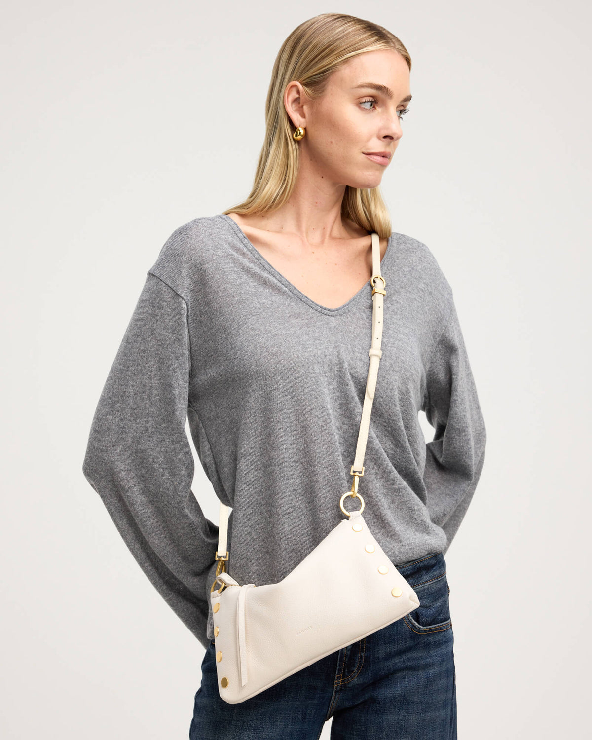A woman wearing a gray long-sleeve V-neck sweater and dark blue jeans, carrying a cream-colored leather shoulder bag with gold hardware and a matching strap.