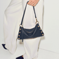 A navy blue leather shoulder bag with gold-tone studs and hardware, held by a model wearing white wide-leg trousers and navy pointed-toe flats against a clean white studio background.
