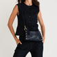A woman wearing a sleeveless black vest and dark pants, carrying a navy blue leather crossbody bag with gold-tone studs and hardware against a light gray studio background.