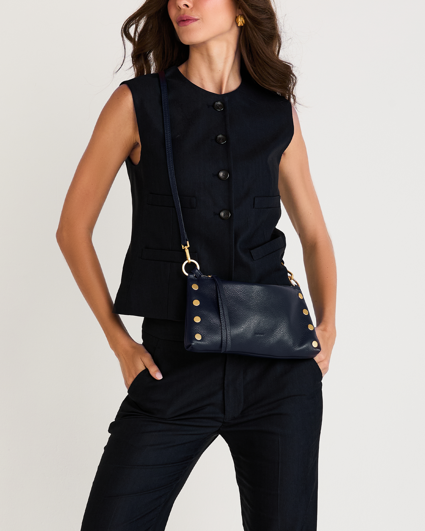 A woman wearing a sleeveless black vest and dark pants, carrying a navy blue leather crossbody bag with gold-tone studs and hardware against a light gray studio background.