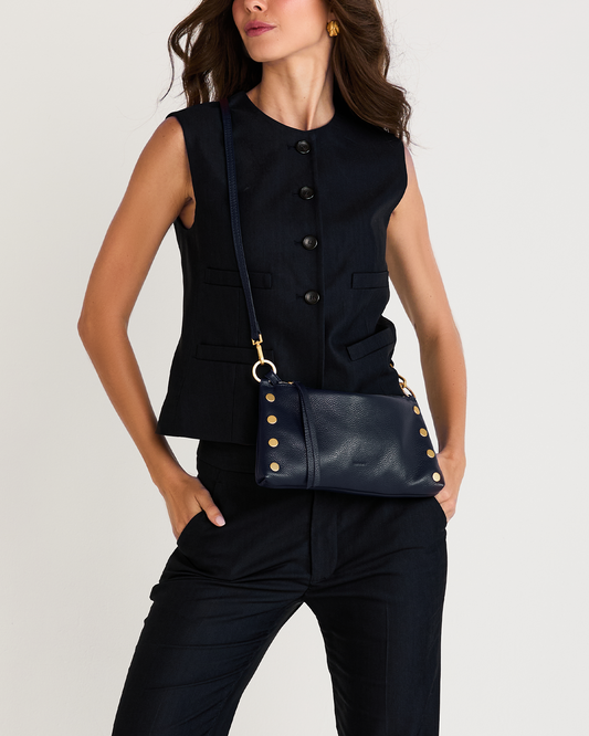 A woman wearing a sleeveless black vest and dark pants, carrying a navy blue leather crossbody bag with gold-tone studs and hardware against a light gray studio background.