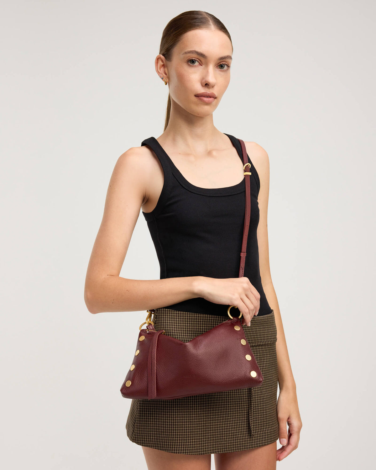 A model wearing a black tank top and a brown houndstooth skirt carries a burgundy leather shoulder bag with gold-tone hardware against a light gray studio background.