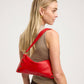 A blonde woman in a beige sleeveless top models a bright red leather shoulder bag with gold-tone hardware and a thin red strap, shown from a side-back profile against a light gray studio background.