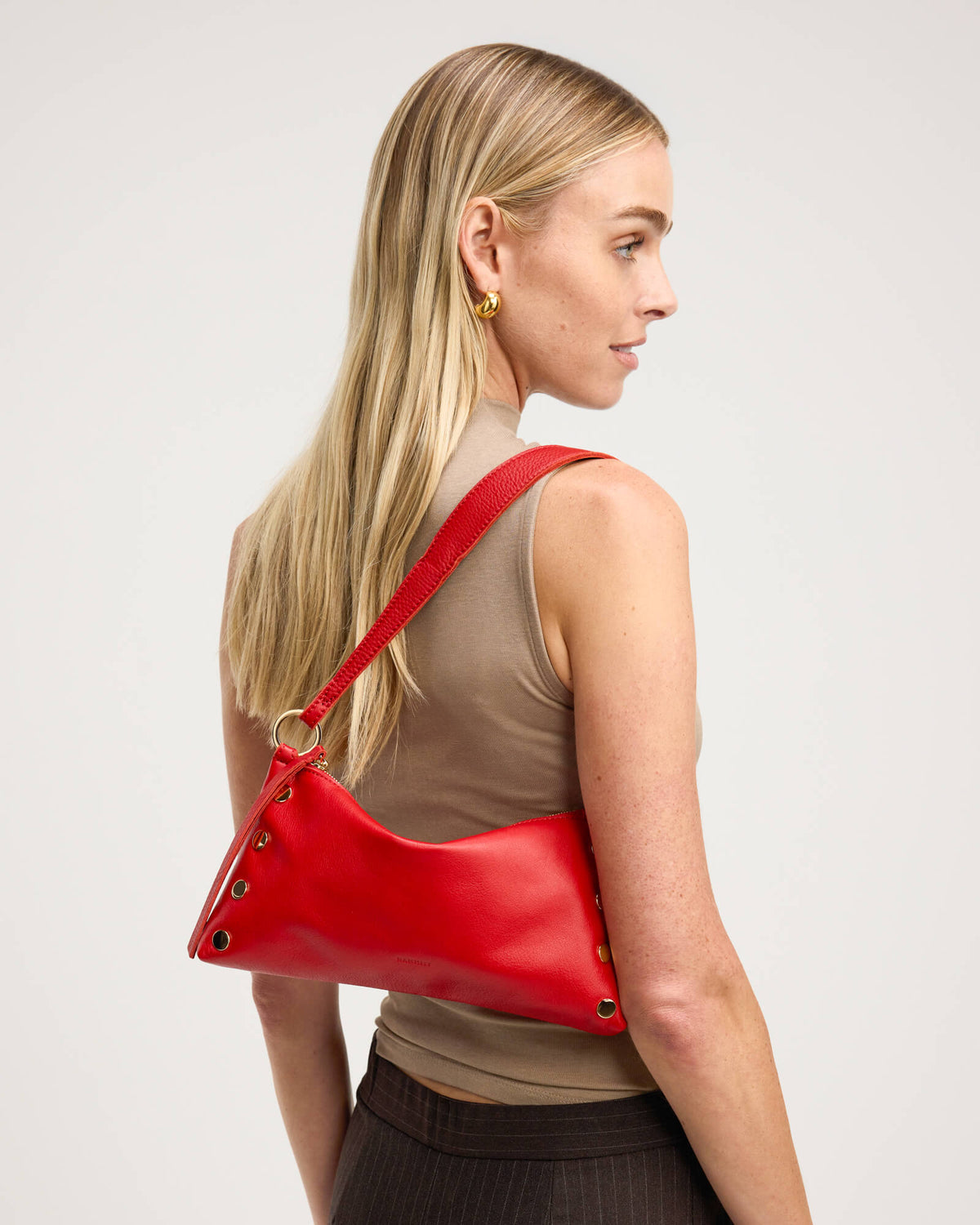 A blonde woman in a beige sleeveless top models a bright red leather shoulder bag with gold-tone hardware and a thin red strap, shown from a side-back profile against a light gray studio background.