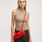 A woman wearing a beige mock-neck top and dark brown pinstriped trousers models a bright red leather shoulder bag with gold hardware against a light gray studio background.