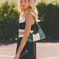 A blonde woman on a tennis court wearing a black and white striped tennis dress and carrying a green and white studded shoulder bag.