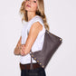 A woman wearing a white sleeveless top and dark jeans carries a large taupe-brown leather shoulder bag with gold-tone studs and a tassel detail against a plain light gray background.