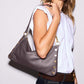 A woman wearing a white sleeveless top and dark blue jeans carries a taupe pebbled leather shoulder bag with gold-tone hardware and snap details against a plain light gray background.